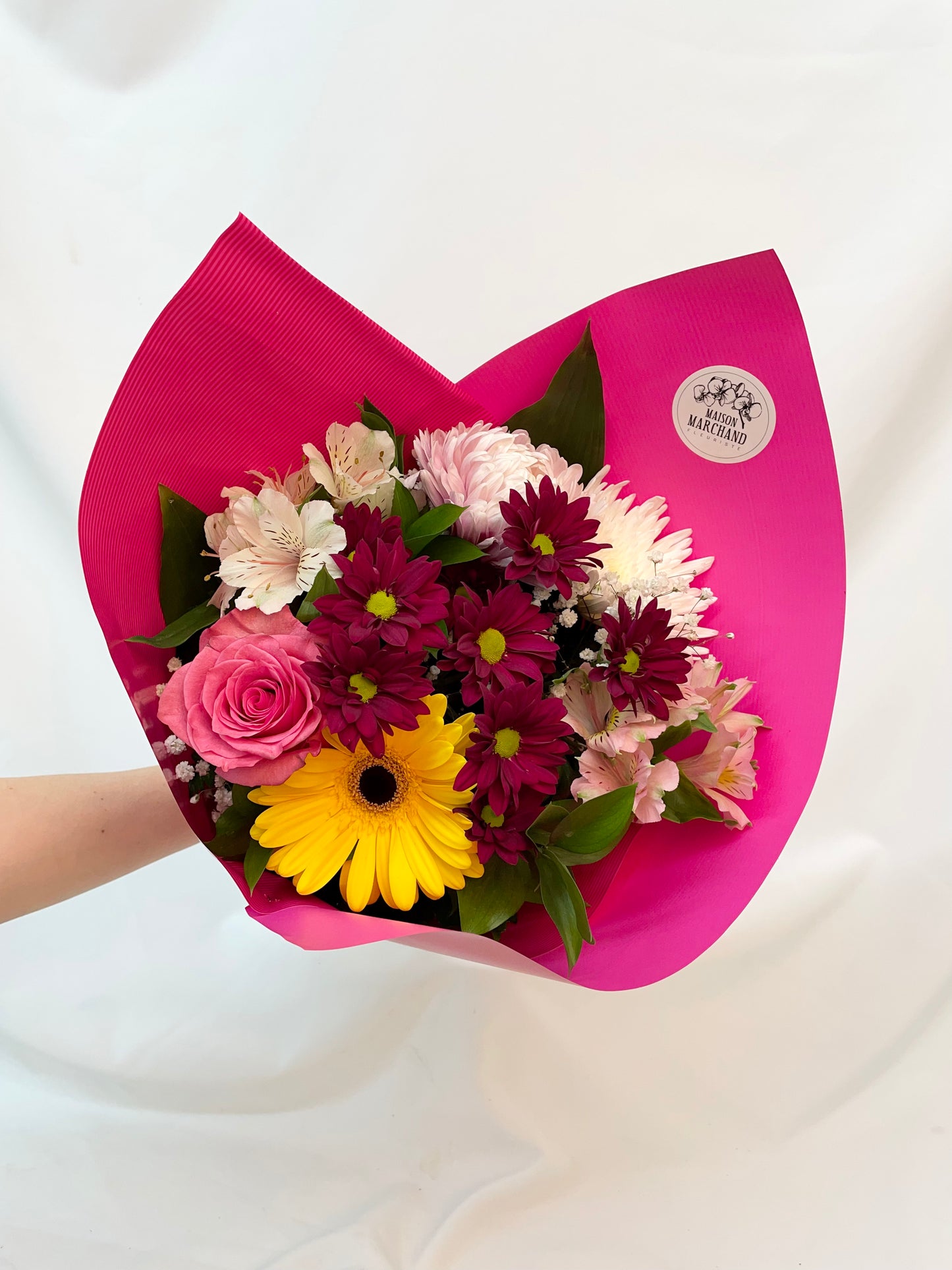 Bouquet “Merci Maman” composé de roses roses, de gerberas jaunes, de chrysanthèmes pourpres, et d’alstroémères blanches, présenté dans un papier rose vif, idéal pour la Fête des Mères.