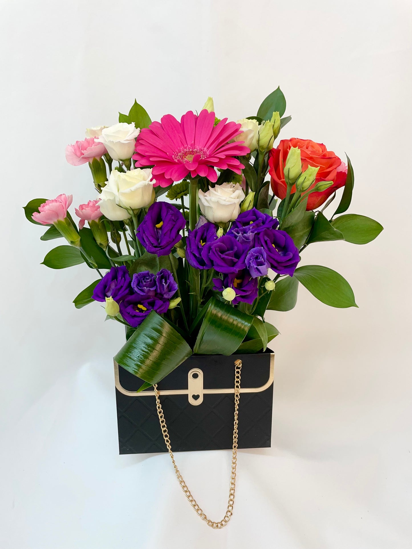 Arrangement floral composé de lisianthus, roses, gerberas et mini œillets, présenté dans un élégant sac à main noir avec une chaîne dorée.