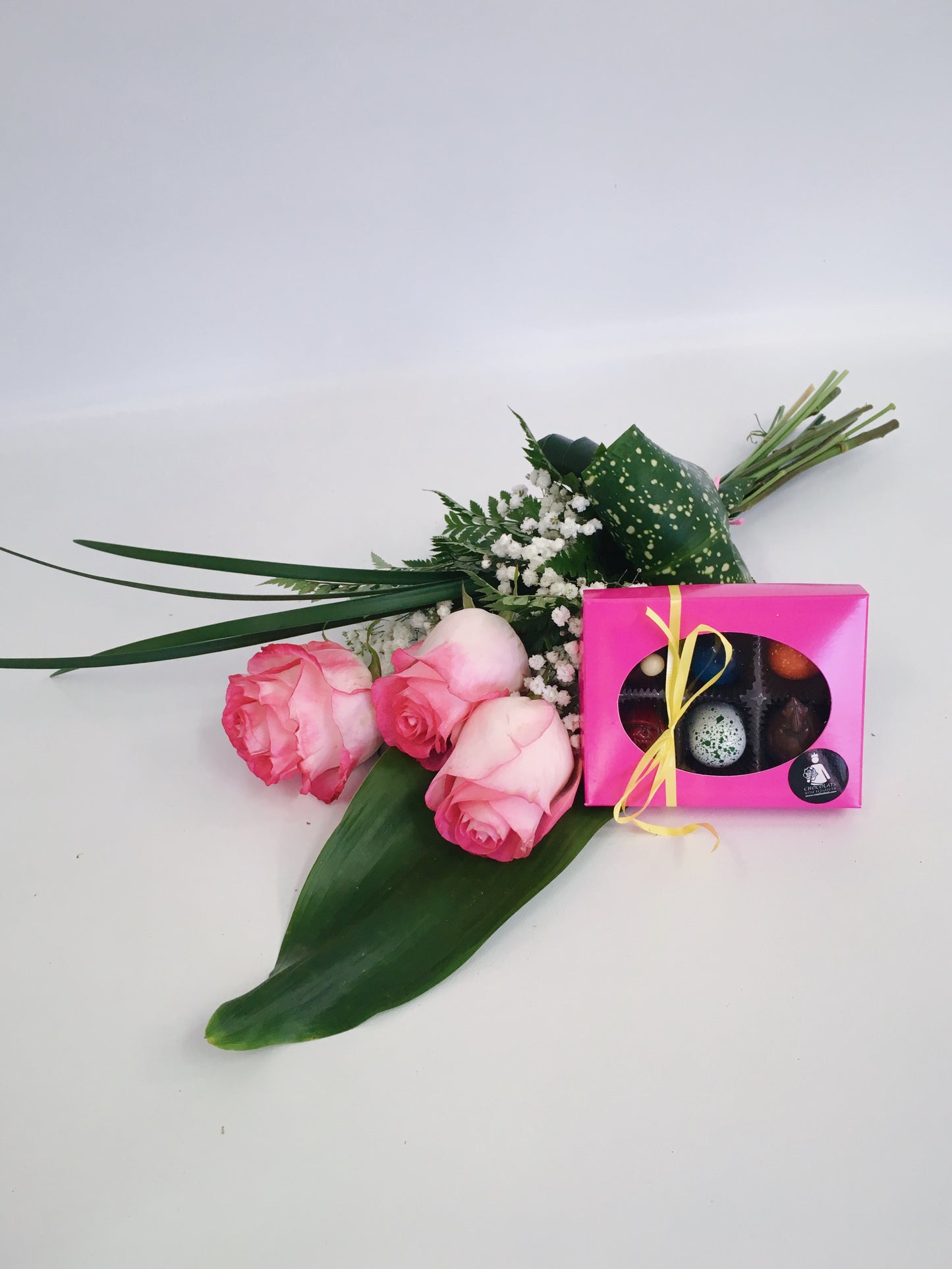 Bouquet de trois roses roses accompagné d’une boîte de chocolats artisanaux emballée dans une boîte rose avec un ruban jaune, le tout disposé sur un fond blanc.