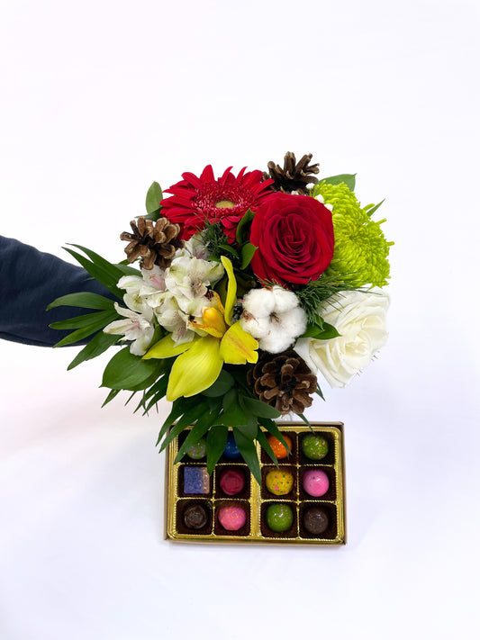 Bouquet de Noël coloré avec roses rouges, gerberas, fleurs jaunes, alstroémères et coton, décoré de cocottes naturelles, présenté avec une boîte de chocolats artisanaux multicolores.