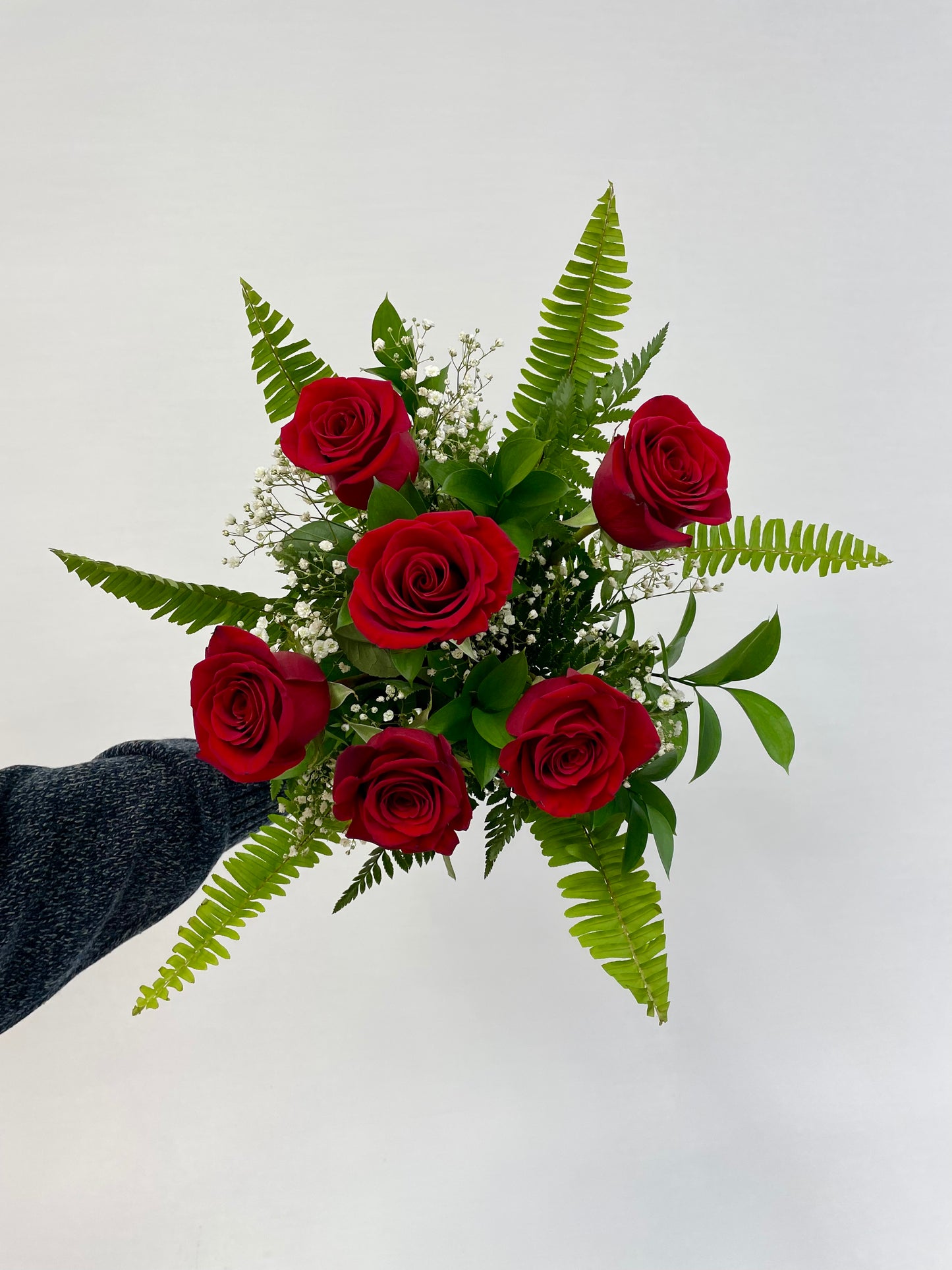 Bouquet de six roses rouges fraîches, entouré de feuillage vert et de gypsophile, tenu par une main contre un fond neutre.