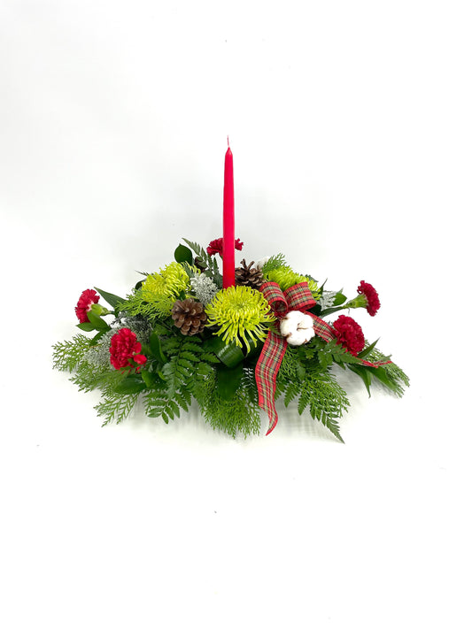 Centre de table festif avec une bougie rouge, des chrysanthèmes verts, des œillets rouges, des pommes de pin, des branches de coton et un ruban écossais.